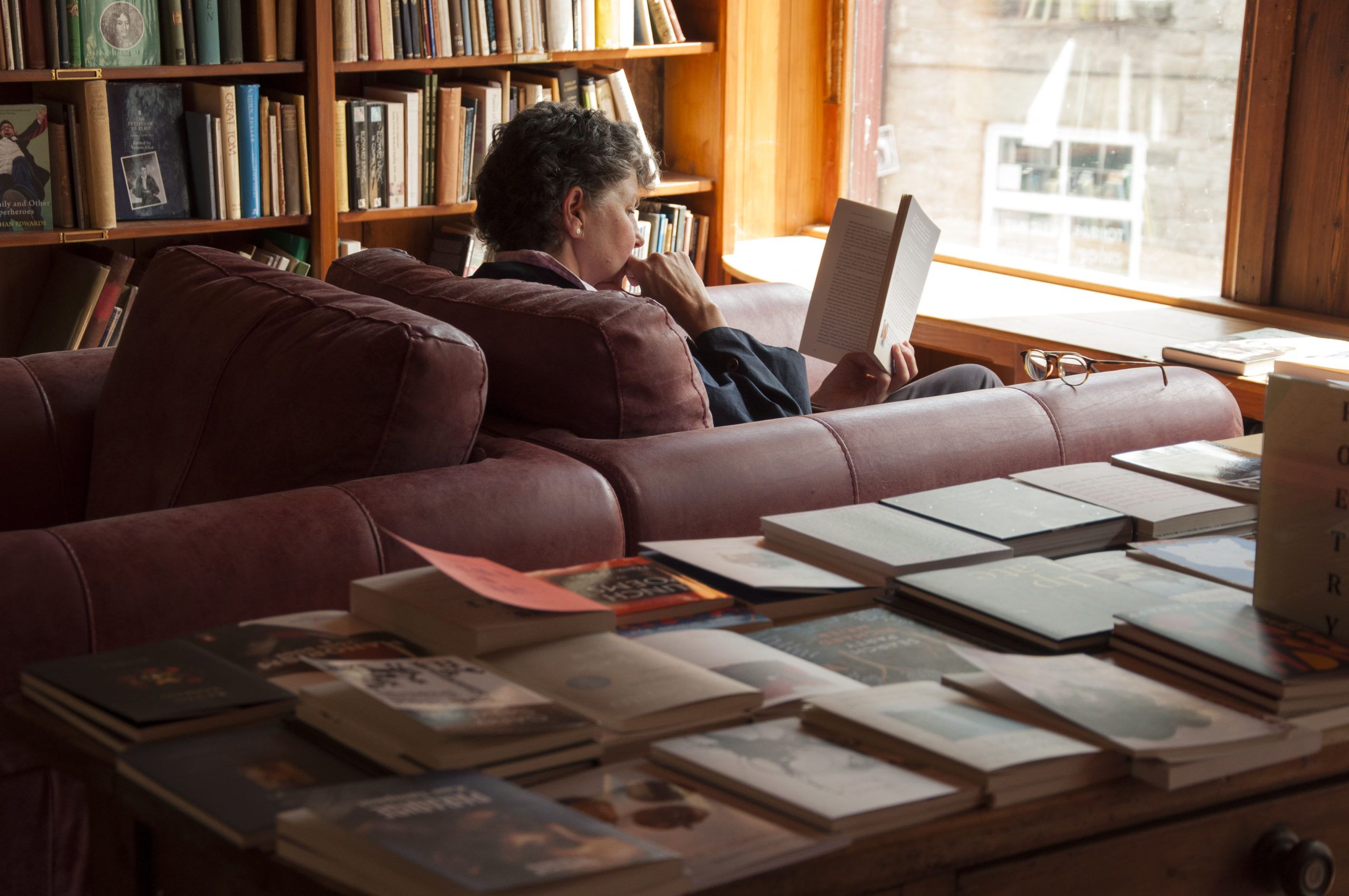 A person is reading a book whilst sat in a chair. At the forefront of the image is a table covered in books that are lying flat