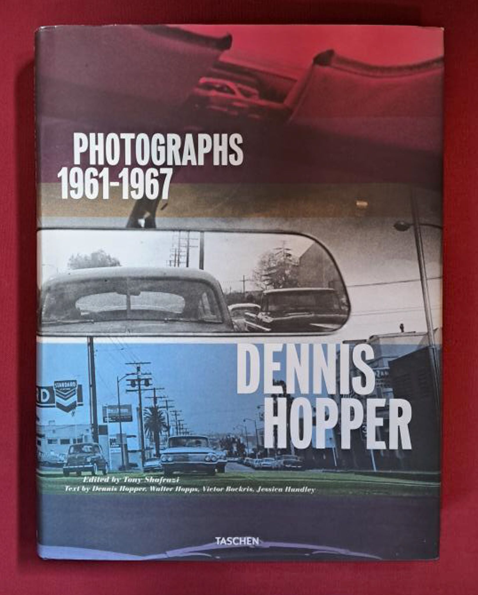 Dennis Hopper: Photographs 1961-1967. The cover Shows a street from the perspective of a driver. In the centre of the image is a rear-view mirror. The image is blue on the bottom third, white in the middle and red at the top. Set on a dark red background